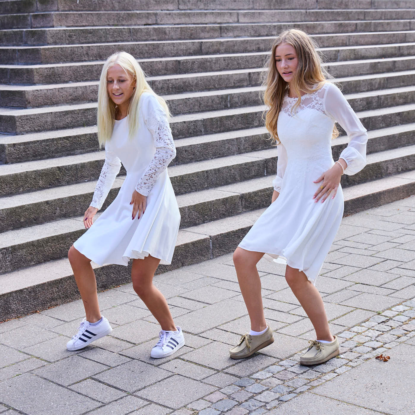 To piger i hvide konfirmationskjoler med lange ærmer og blonder, danser foran en trappe